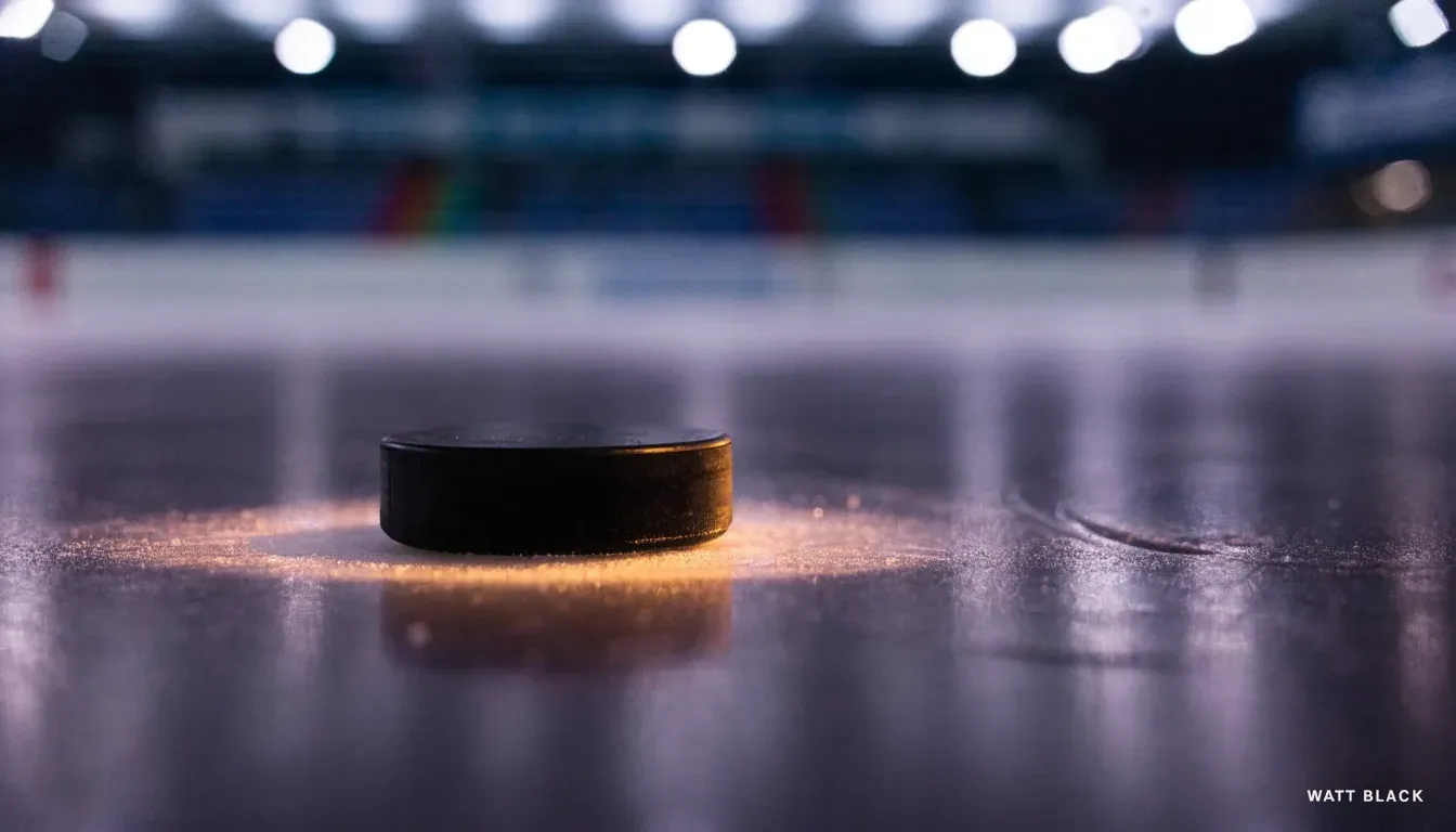 Ein Eishockeypuck liegt auf dem Eis, im Hintergrund unscharf die Bande unter hellem Hallenlicht.