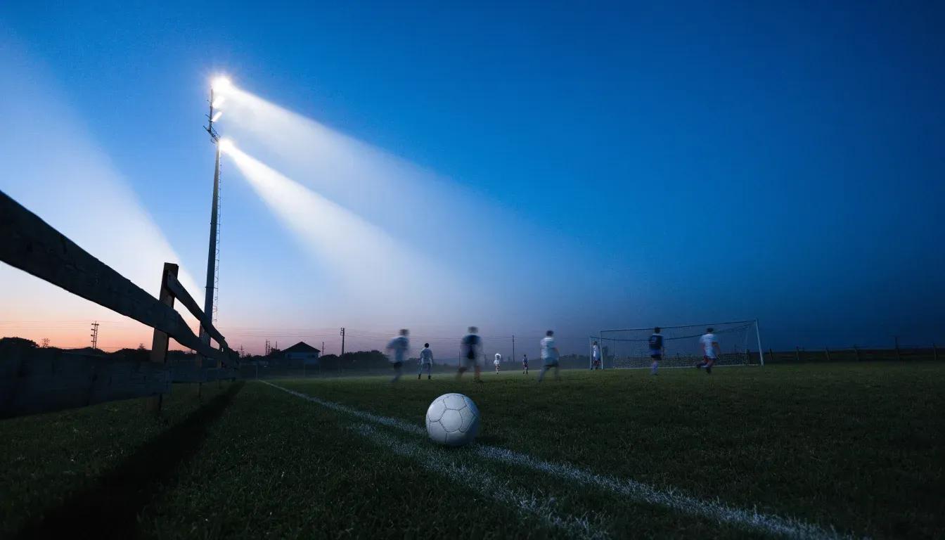 Ein kleiner Dorf-Fussballplatz liegt in der Dämmerung unter einem hohen Flutlichtmast, am Spielfeldrand steht ein Holzzaun.