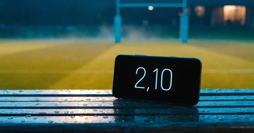 Ein ovaler Rugbyball liegt auf feuchtem Rasen vor zwei hohen Torstangen, hinter dem Spielfeld steigen Nebelschwaden auf.