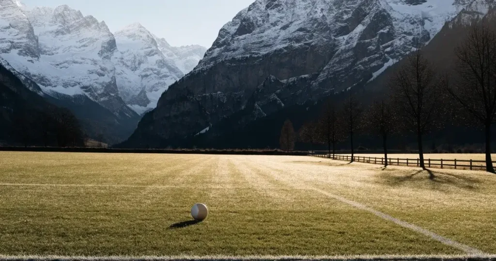 Ein Dorf-Sportplatz mit einem Ball am Strafraumrand, dahinter schneebedeckte Alpengipfel im klaren Morgenlicht