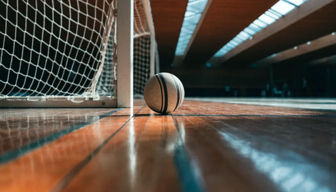 Ein Handball liegt am Rand des Torraums in einer leeren Sporthalle, Licht fällt durch Oberlichter auf den Hallenboden.